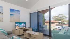 Ein modernes Wohnzimmer mit Fernseher, Schreibtisch und einer Couch. Ein Schiebeglasfenster führt zu einem Außenbereich mit Blick auf die Nachbarschaft.