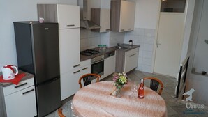 Een kleine keuken met een koelkast, fornuis en wastafel. Er staat een eettafel met stoelen en een vaas met bloemen.
