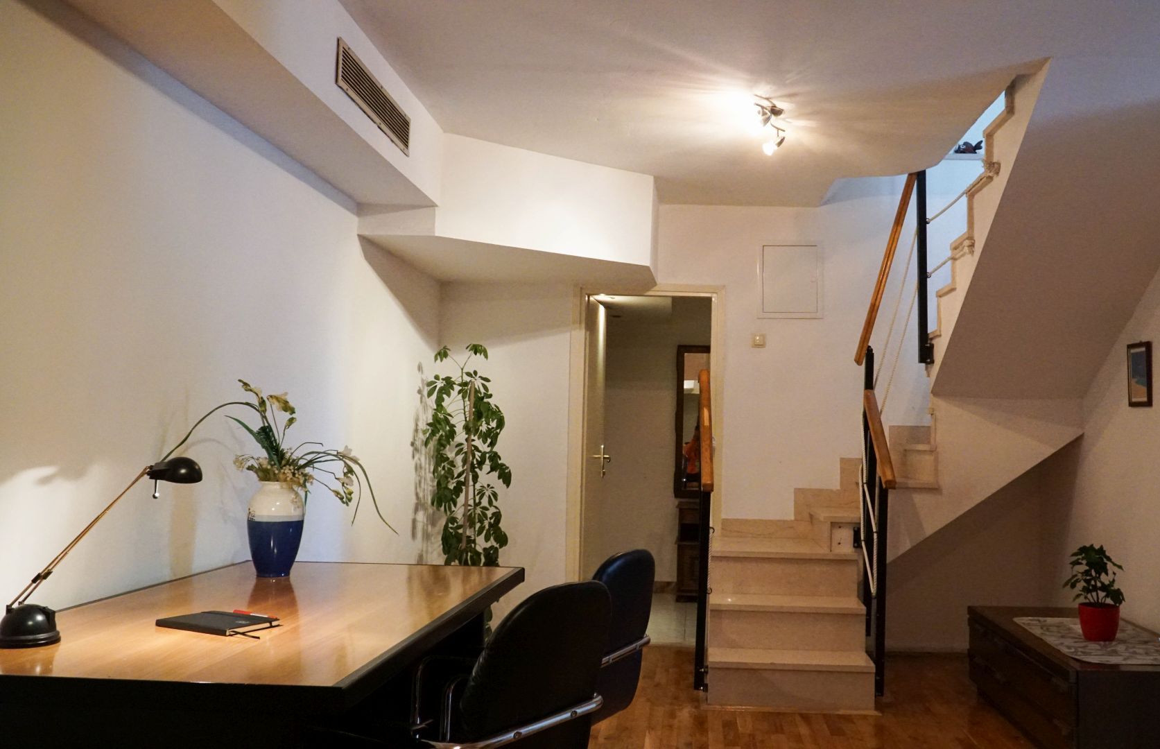 A modern living room with a wooden dining table, two chairs, a desk with a lamp, and a potted plant. A staircase leads to an upper floor.