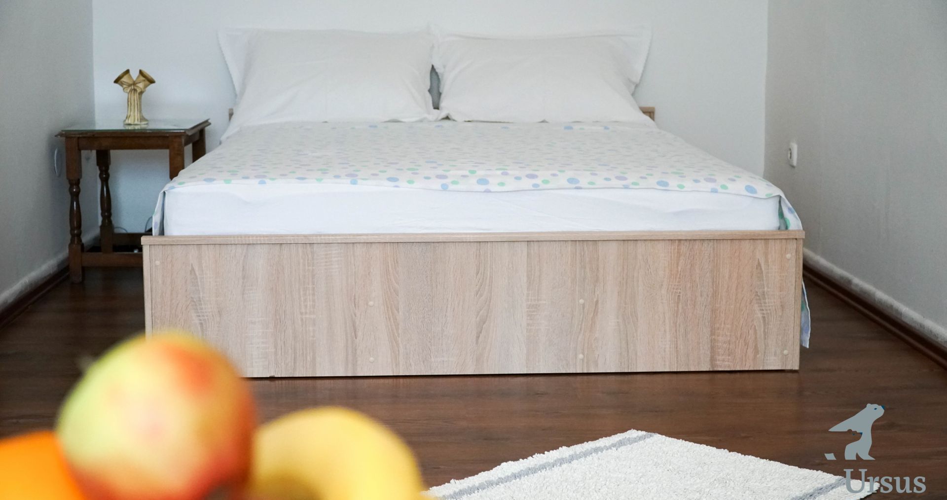 A bedroom with a wooden bed frame, white bedding, a small bedside table with a vase, and a fruit bowl on the floor.