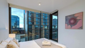 Une chambre d’hôtel moderne dotée d’un grand lit, offrant une vue sur le paysage urbain grâce à de larges fenêtres du sol au plafond, et agrémentée d’une œuvre d’art contemporaine au mur.
