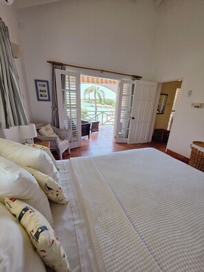 Ein Schlafzimmer mit einem großen Bett, einem Sessel, einem Tisch und Blick auf einen Pool und Palmen durch offene Türen.