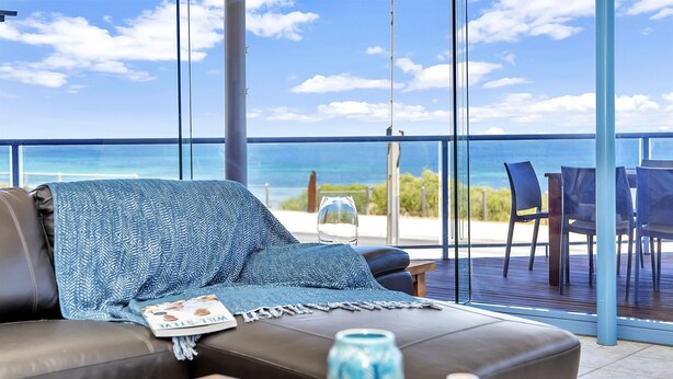 A balcony with a view of the sea, a leather sofa with a blue blanket, and a table with a blue vase. - Sail Away - Aldinga Beach - C21 SouthCoast Holidays (Aldinga Beach)