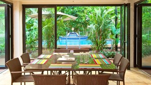 A dining area with a glass table, wicker chairs, and a view of a pool and tropical plants. - 7 Solander Boulevard, Port Douglas QLD 4877 (Port Douglas)