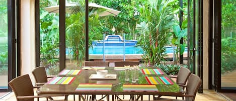 A dining area with a glass table, wicker chairs, and a view of a pool and tropical plants.