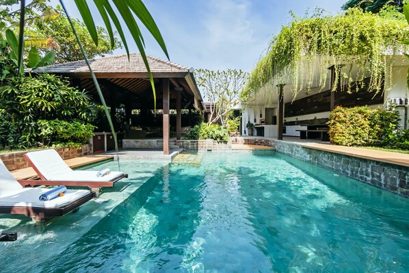 A swimming pool with lounge chairs, a covered seating area, and lush greenery.