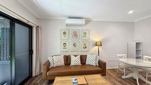 Una sala moderna con un sofá de cuero marrón, una mesa de centro de madera y una mesa de comedor de vidrio con sillas blancas. Hay cuadros enmarcados en la pared y una lámpara de pie.
