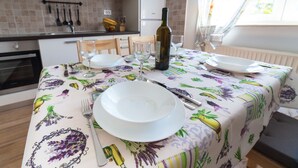 A dining table set with white plates, a wine bottle, and cutlery in a kitchen with a tiled backsplash and a window with curtains. - Apartment Raspolic - no.1 (Trinajstici)