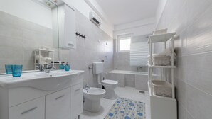 A modern bathroom with a white sink, bidet, toilet, and bathtub. There are white shelves with towels and a blue patterned rug. - Apartment Raspolic - no.1 (Trinajstici)