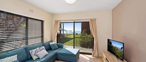 A living room with a blue sofa, a TV, and a balcony with a view of the sea.