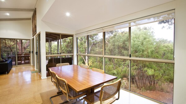 A spacious dining area with a large wooden table and woven chairs, large windows offering a view of a forested landscape.