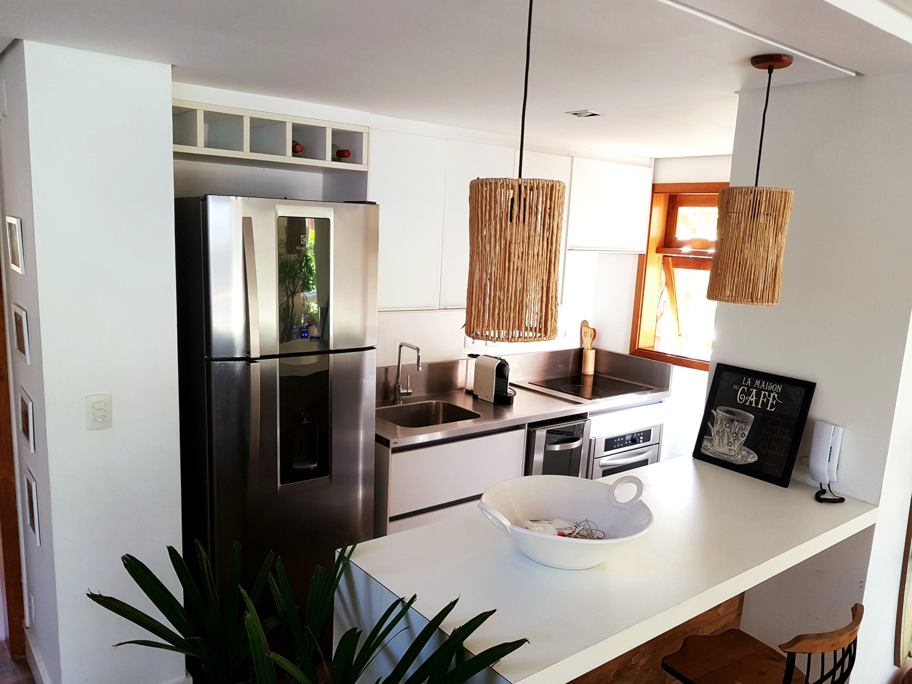 Una cocina moderna con un refrigerador de acero inoxidable, un lavamanos y una encimera blanca. Hay dos lámparas colgantes y un menú enmarcado en la pared.