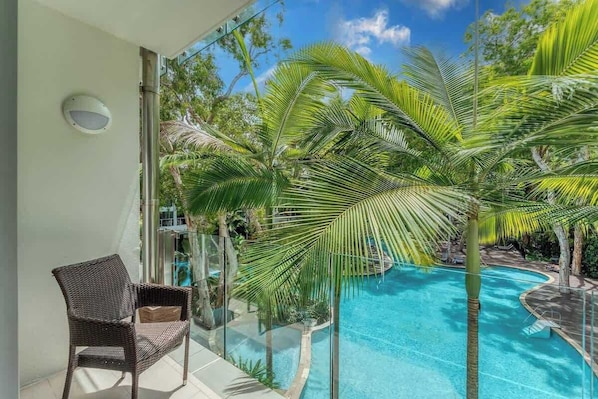Un balcone con una sedia in vimini, una ringhiera in vetro e vista su una piscina circondata da palme.