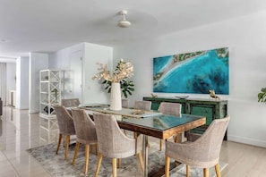 A dining area with a glass-top table, wicker chairs, a white vase with flowers, a green cabinet, and a large abstract painting on the wall.