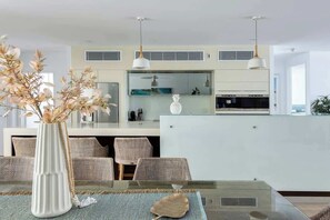 Una cucina moderna con isola centrale, illuminazione a sospensione e zona pranzo con tavolo in vetro e sedie in vimini.