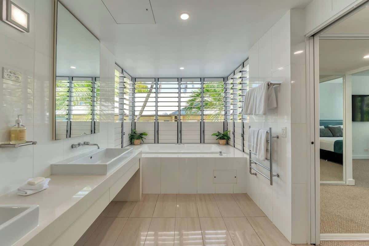 Ein modernes Badezimmer mit einer großzügigen Badewanne, zwei Waschbecken, einem Spiegel und einem Fenster mit Jalousien.