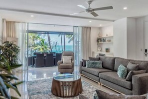 Un soggiorno moderno con divano, poltrona e tavolino da caffè, una ventola a soffitto e vista sull'oceano attraverso ampie finestre.