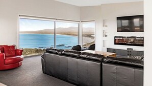A modern living room with a large black leather sofa, a red armchair, and a kitchen area with a flat-screen TV. - Tranquility Bay of Fires - A beautiful beach house in a stunning location (The Gardens)