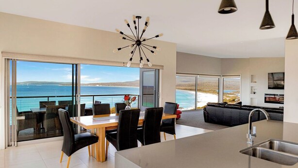 A modern kitchen with a large island, a dining area with a view of the sea, and a living area with a sofa and a TV. - Tranquility Bay of Fires - A beautiful beach house in a stunning location (The Gardens)