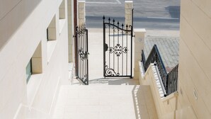 Un escalier surmonté d’une grille en fer forgé, menant à un bâtiment doté d’un sol carrelé et de piliers en pierre.