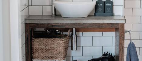 Un tocador de baño con un lavabo blanco, dos botellas de aceite de oliva, una cesta tejida y un perchero para toallas.