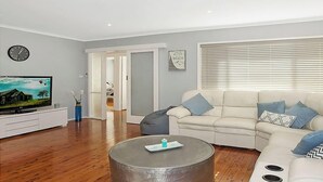 Un soggiorno moderno con TV a schermo piatto, un divano bianco con cuscini blu, un tavolino rotondo e pavimento in legno.