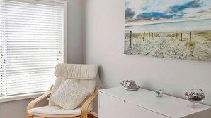 Una stanza con una sedia bianca, un comò bianco, un quadro che raffigura una spiaggia e una finestra con persiane.
