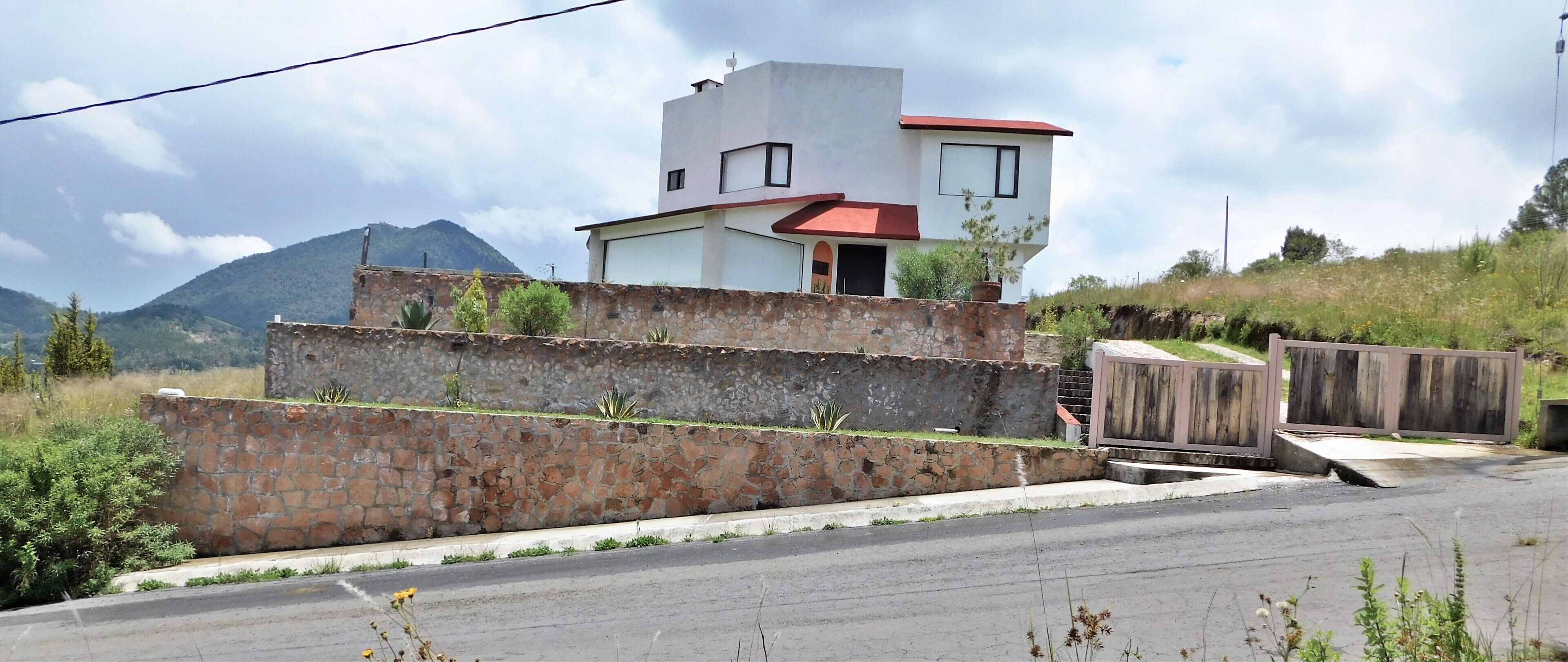 Un bâtiment moderne à deux étages, doté d’un toit rouge, d’un mur de soutènement en pierre et offrant une vue sur une montagne en arrière-plan.