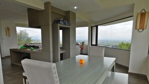 Un espace repas moderne avec une grande table et des chaises, un coin cuisine doté d’un four encastré et de grandes fenêtres offrant une vue sur la campagne.