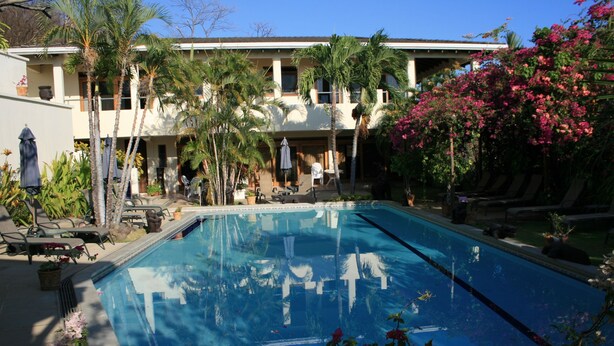 A swimming pool with lounge chairs and umbrellas, surrounded by palm trees and a building with a balcony. - Villas Casa Loma - Suite 203 : Tropical Villa with Pool and Spectacular Views (Playa Flamingo)