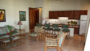 A living room with a sofa, coffee table, and chairs, a kitchen with wooden cabinets and appliances, and a door leading to another room. - Villas Casa Loma - Suite 203 : Tropical Villa with Pool and Spectacular Views (Playa Flamingo)