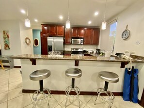 Una cocina moderna con armarios de madera, electrodomésticos de acero inoxidable y encimera de granito. Hay tres taburetes de bar y un colgante.