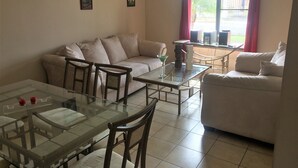 A living room with a sofa, a glass coffee table, and a dining table with chairs. There are three framed pictures on the wall.