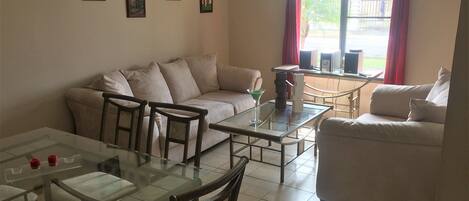 A living room with a sofa, a glass coffee table, and a dining table with chairs. There are three framed pictures on the wall.