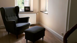 A room with a dark armchair and ottoman, a radiator, a window with a view of greenery, and a framed picture on the wall.