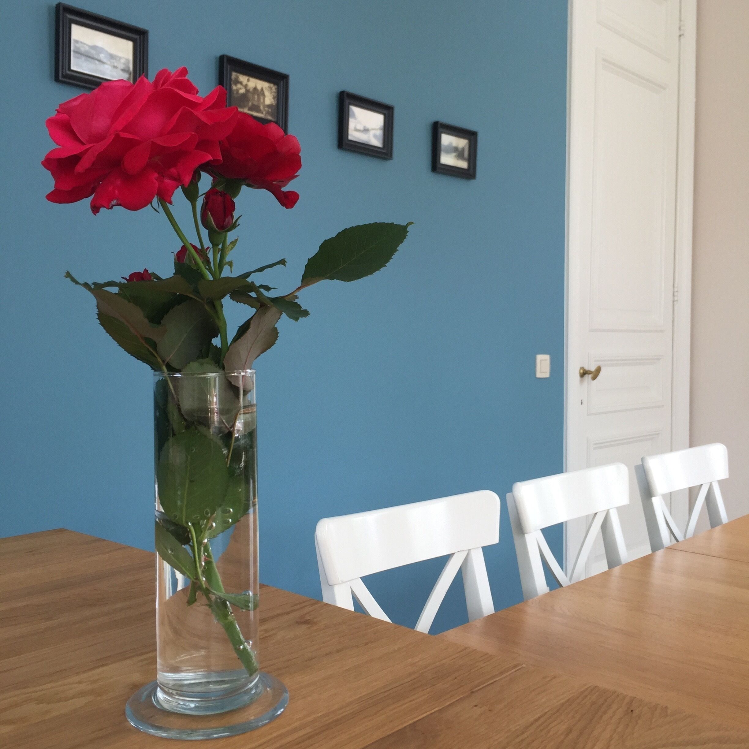 A clear glass vase with red roses on a wooden table against a blue wall with framed pictures.