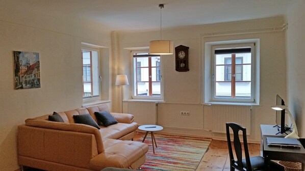 A living room with a sofa, a small round table, and a chair. There are two windows with curtains, a radiator, and a clock on the wall. - Jager 2 Tour As / 1-room APP with AC, Wi-Fi, in the pedestrian zone (Ljubljana)