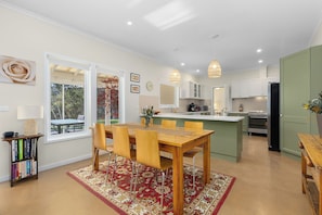 A modern kitchen with white cabinets, a dining area with a wooden table and chairs, and a view of the outdoors through large windows.