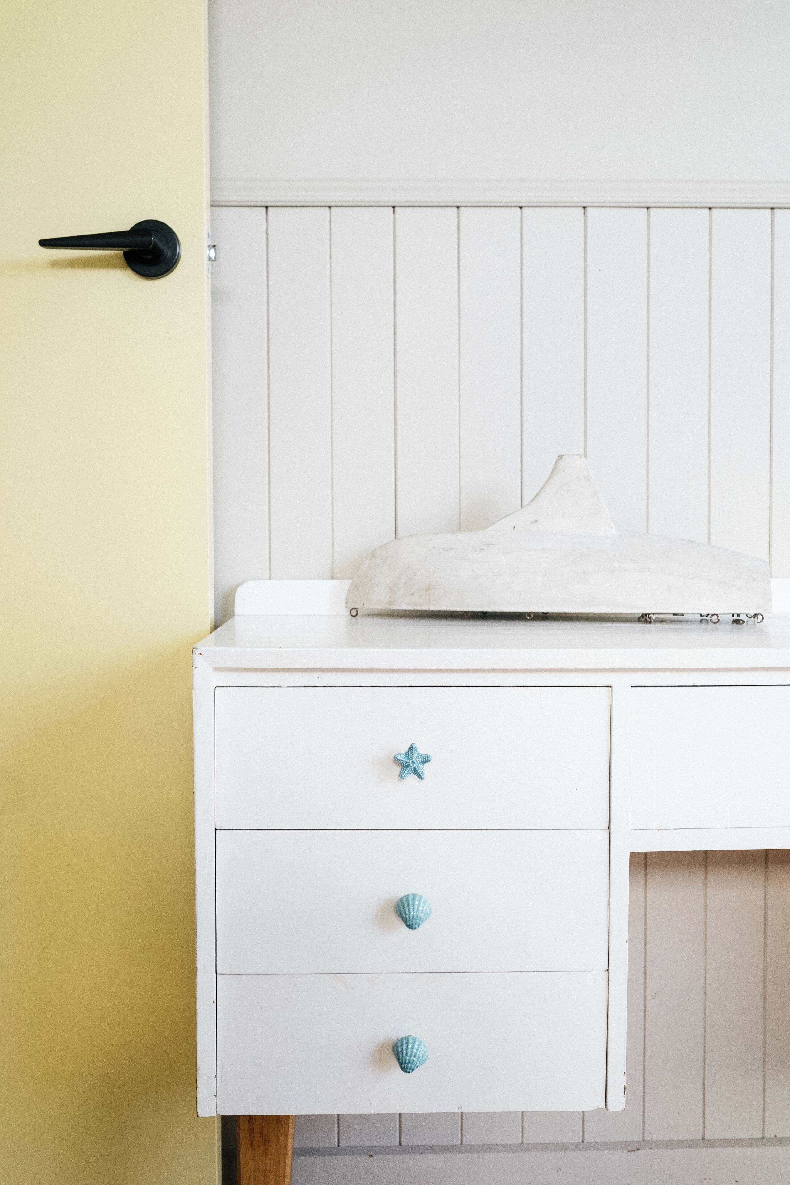 Un aparador blanco con un tirador en forma de estrella de mar, un mueble blanco con tapa de mármol y una puerta amarilla.