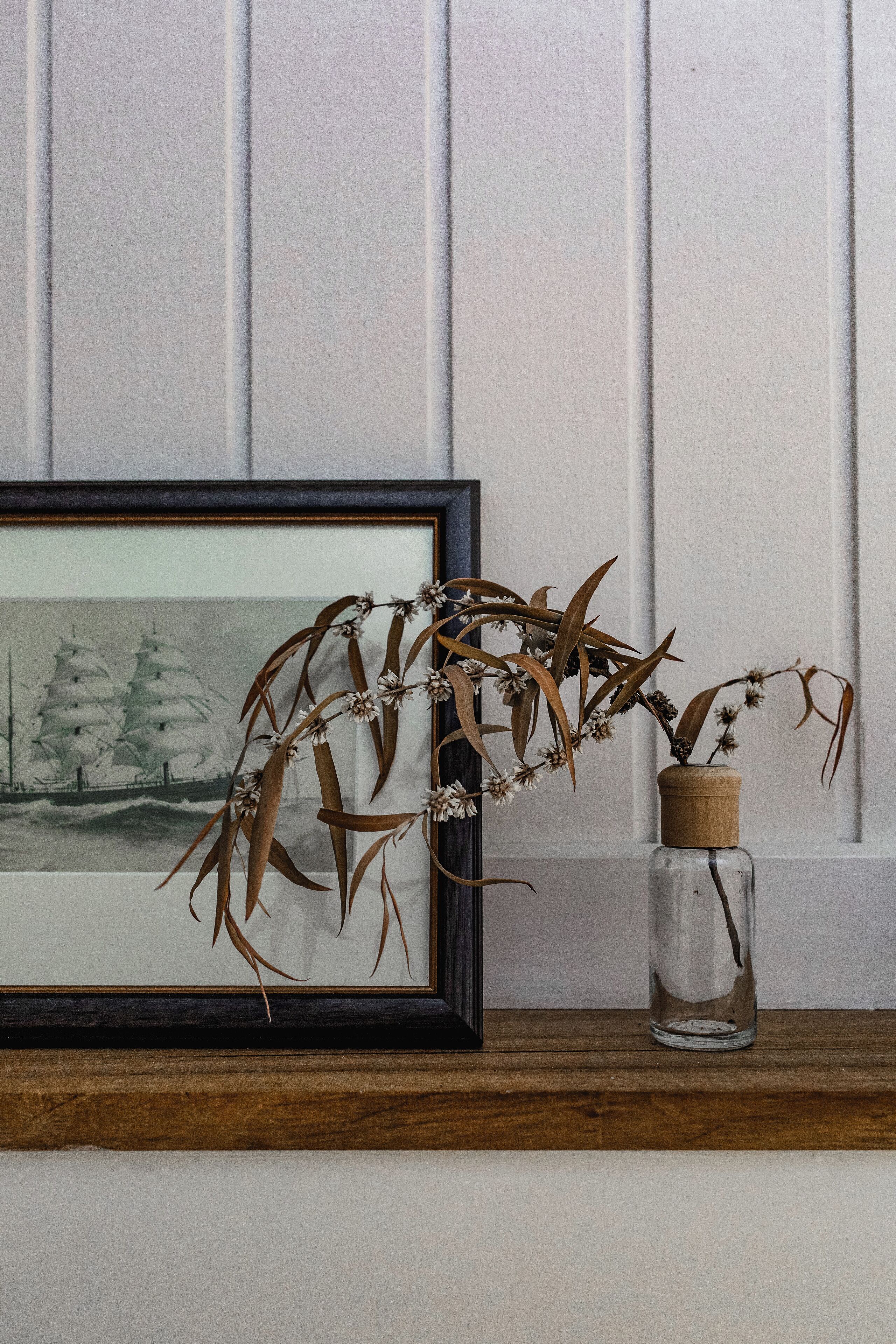A framed artwork of sailboats on a shelf, a clear glass vase with dried plants, and a wooden shelf against a white paneled wall.
