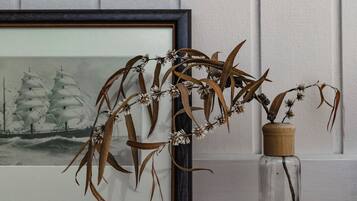 A framed artwork of sailboats on a shelf, a clear glass vase with dried plants, and a wooden shelf against a white paneled wall.