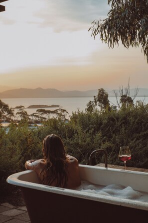 A bathtub with a glass of red wine, surrounded by greenery and overlooking a body of water with distant landforms. - Relax, Restore and Explore at The Sandy Sister (Dodges Ferry)