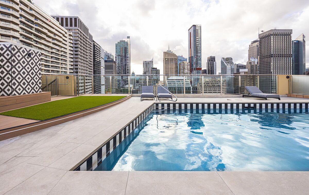 Piscina no terraço com duas espreguiçadeiras, guarda-corpo de vidro e paisagem urbana ao fundo.