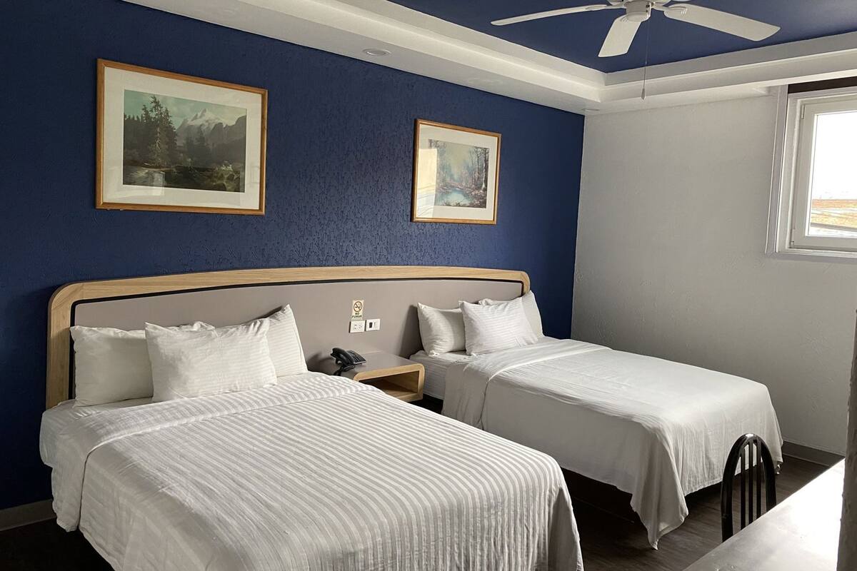 Habitación de hotel con dos camas, ventilador de techo, fotografías enmarcadas en la pared y ventana con vistas.