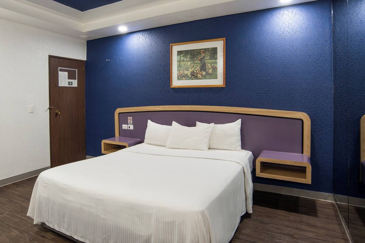 Habitación de hotel con cama, dos mesitas de noche, cabecera de madera, un cuadro en la pared y una puerta con un letrero.