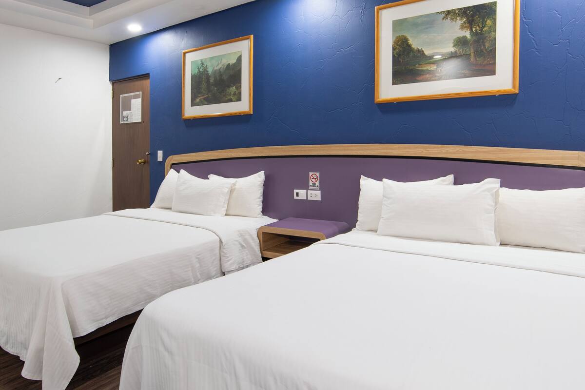 Habitación de hotel con dos camas individuales, cabecero de madera, pared de acento morada y dos cuadros con paisajes.