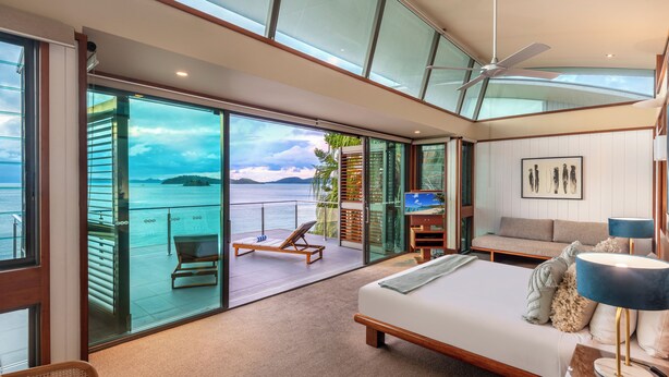 Ein modernes Schlafzimmer mit einem großen Bett, einem Schreibtisch und einem Stuhl. Es verfügt über einen Balkon mit Meerblick und eine Sitzecke mit einem Sessel und einem Tisch.