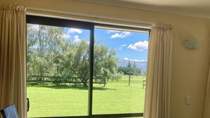 Una habitación con un amplio ventanal corredero que da a un área exterior con césped, un escritorio de madera con una silla y un sofá con una manta.