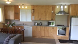 Cocina compacta con armarios de madera, lavavajillas, microondas, estufa y refrigerador. Hay un comedor con mesa y sillas, y un sofá en la sala.