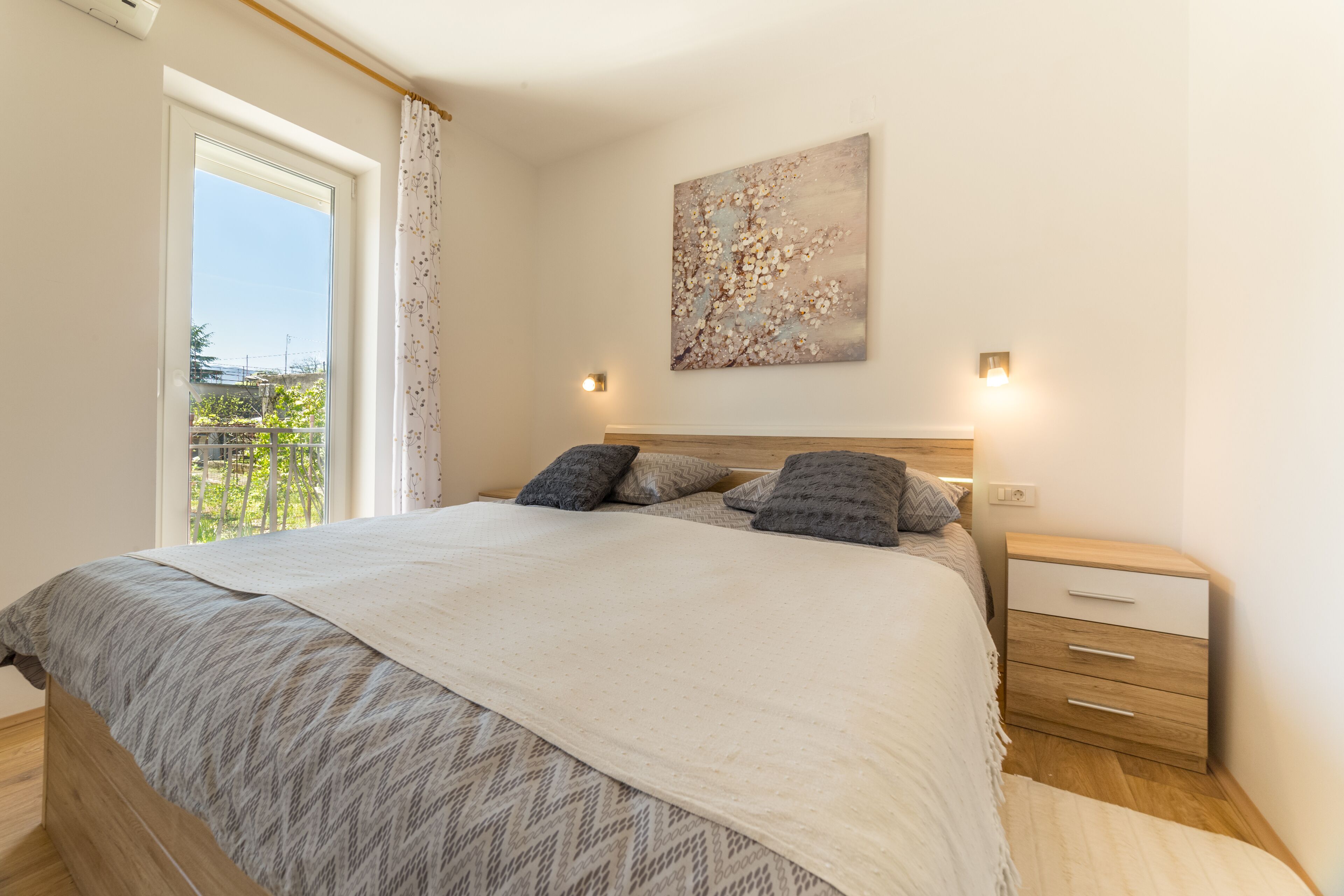 Een slaapkamer met een groot bed, een nachtkastje met lades, een wandlamp en een raam met gordijnen.